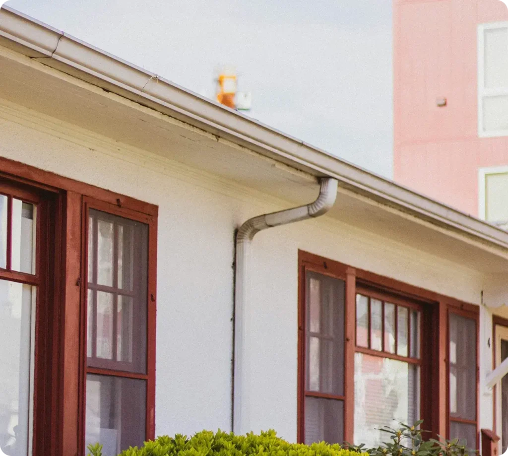 Vit byggnad med röda inramade fönster och ränna, omgiven av grönska och andra byggnader.