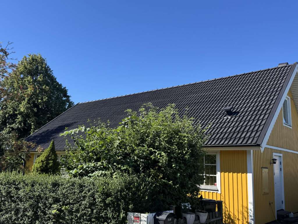 Gult hus med svart tegeltak, takmålning Skåne, och grönska under en klarblå himmel.