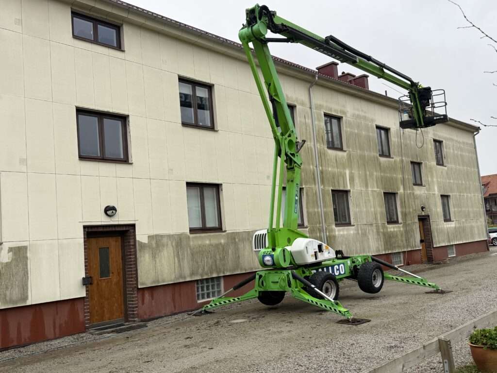 Grön skylift som står framför en smutsig lägenhetsbyggnad. Byggnaden håller på att högtrycksspolas och halva fasaden är ren och fin.