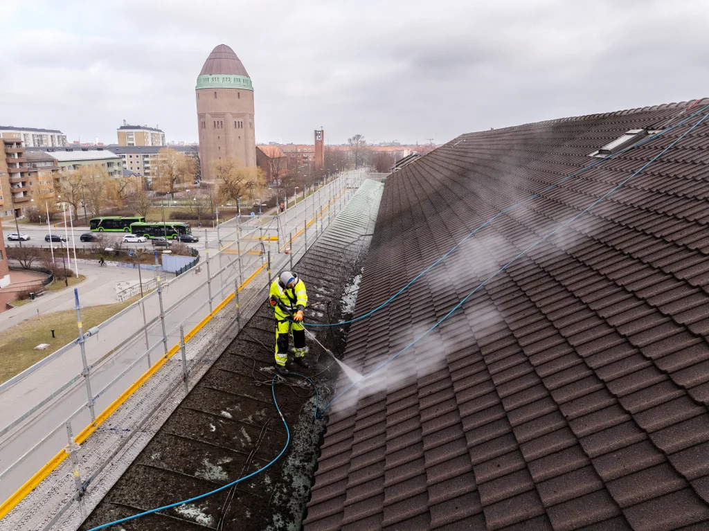 En drönarbild som visar professionell taktvätt i Malmö, Skåne, utförd vid Södervärn. En taktvättare i varselkläder använder högtryckstvätt på ett stort tegeltak med Södervärns karakteristiska vattentorn och stadstrafik i bakgrunden.